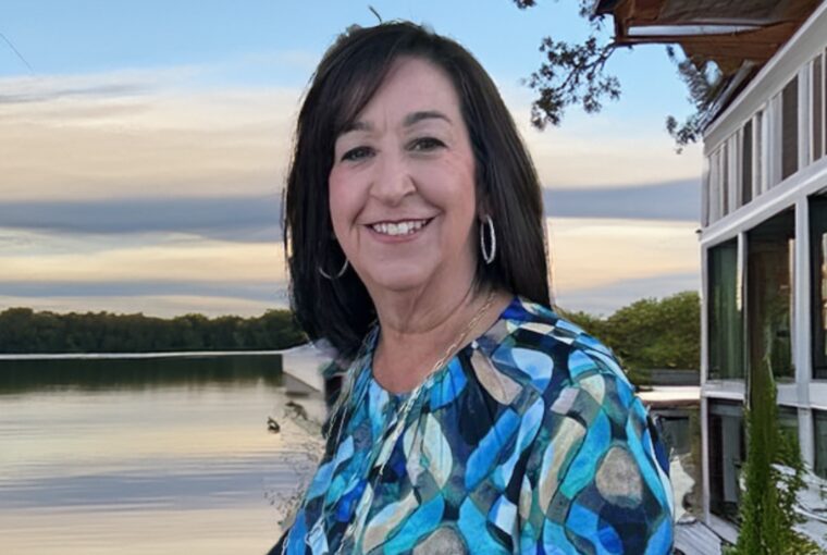 A woman in a red top is standing in front of a serene, wooded lakeside background.