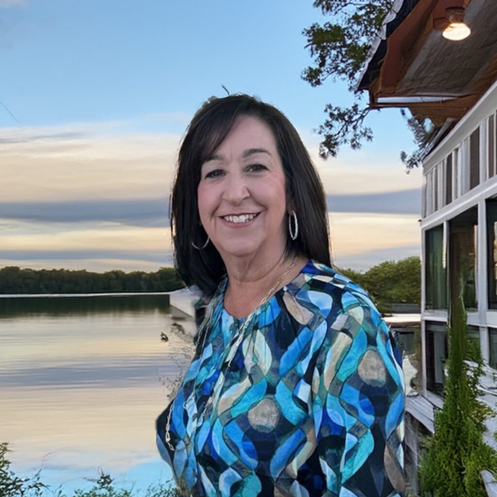 A woman wearing a red blouse stands smiling in front of a scenic lake view with houses and trees in the background.