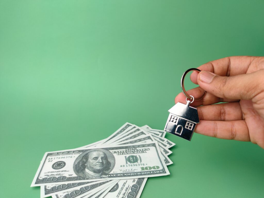 A hand holds a house-shaped keychain above a stack of $100 bills against a green background, capturing the dream of owning a lakeside property at Lake Martin.