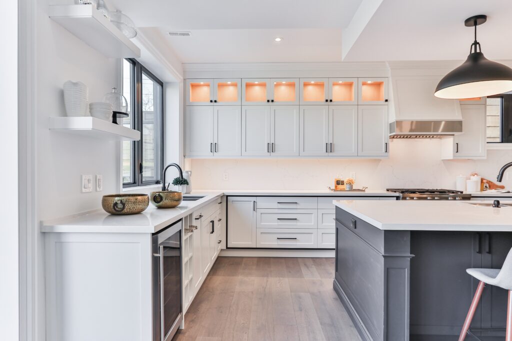 Modern kitchen with white cabinets, light wood flooring, a large island with a dark base, stainless steel appliances, and under-cabinet lighting. There's a window over the sink offering views of Lake Martin and a pendant light above the island.