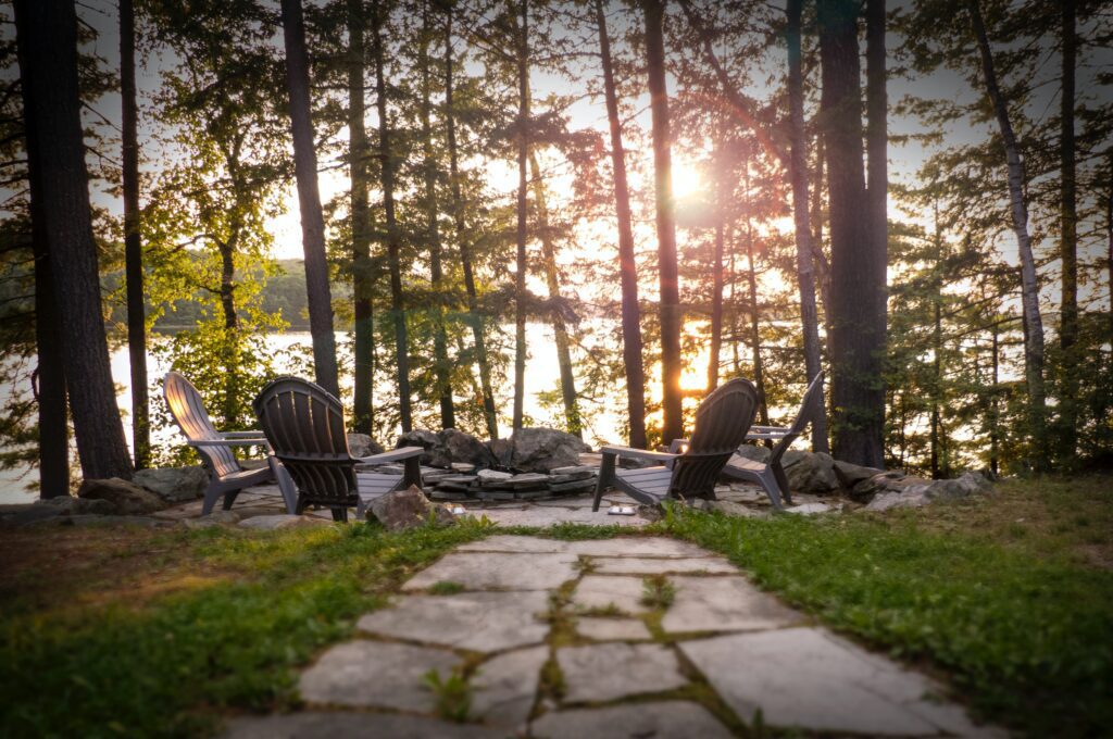 Four Adirondack chairs encircle a stone fire pit in a wooded area near Lake Martin, Alabama, with a path leading up to them. The sun is setting behind the trees and casting a warm glow over the scene.