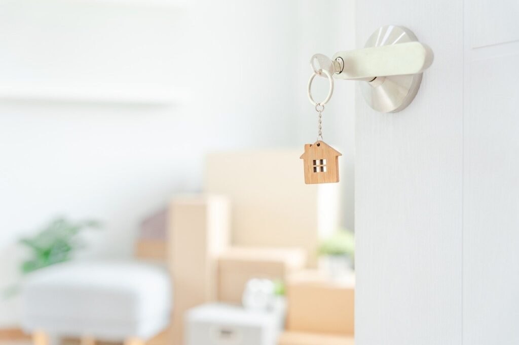 A wooden house-shaped keychain is inserted in a door lock. Blurred moving boxes and furniture are visible in the background, suggesting a newly occupied Lake Martin home or one soon to be.