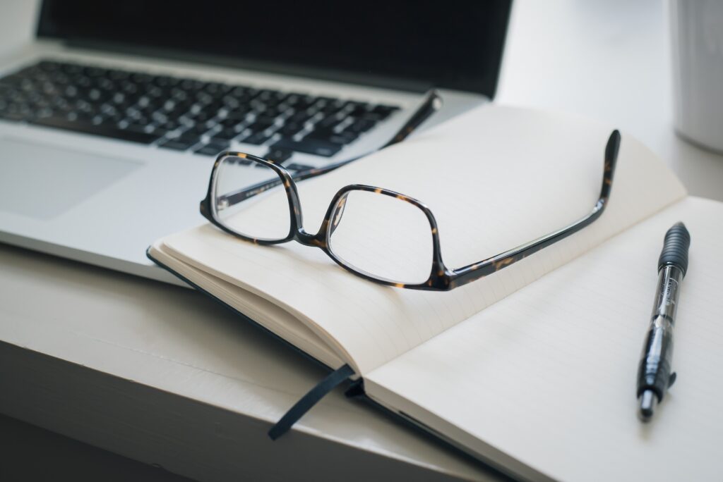 A pair of eyeglasses rests on an open notebook next to a pen and a laptop on a desk, with a picturesque photo of Lake Martin propped up beside them.