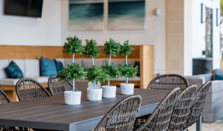 A modern dining area features a long table with six small potted plants as a centerpiece, surrounded by wicker chairs, evoking the serene vibes of Lake Martin. In the background, a cozy couch and tasteful wall art complete the inviting atmosphere.