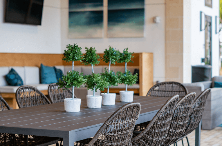 A modern dining area features a long table with six small potted plants as a centerpiece, surrounded by wicker chairs, evoking the serene vibes of Lake Martin. In the background, a cozy couch and tasteful wall art complete the inviting atmosphere.