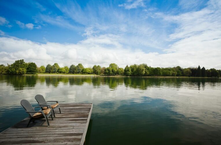 Two empty wooden chairs on a dock overlook a calm, reflective lake surrounded by lush, green trees under a partly cloudy sky.