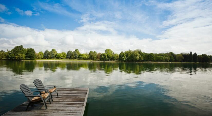 Two empty wooden chairs on a dock overlook a calm, reflective lake surrounded by lush, green trees under a partly cloudy sky.