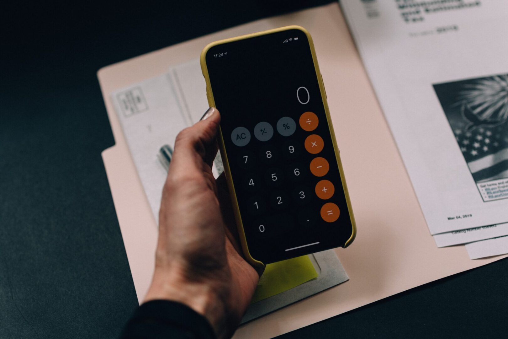 A hand holding a smartphone with a calculator app open, displaying zero. Background includes paperwork, partially shown, on a dark surface, perhaps calculating the budget for a potential Lake Martin home.